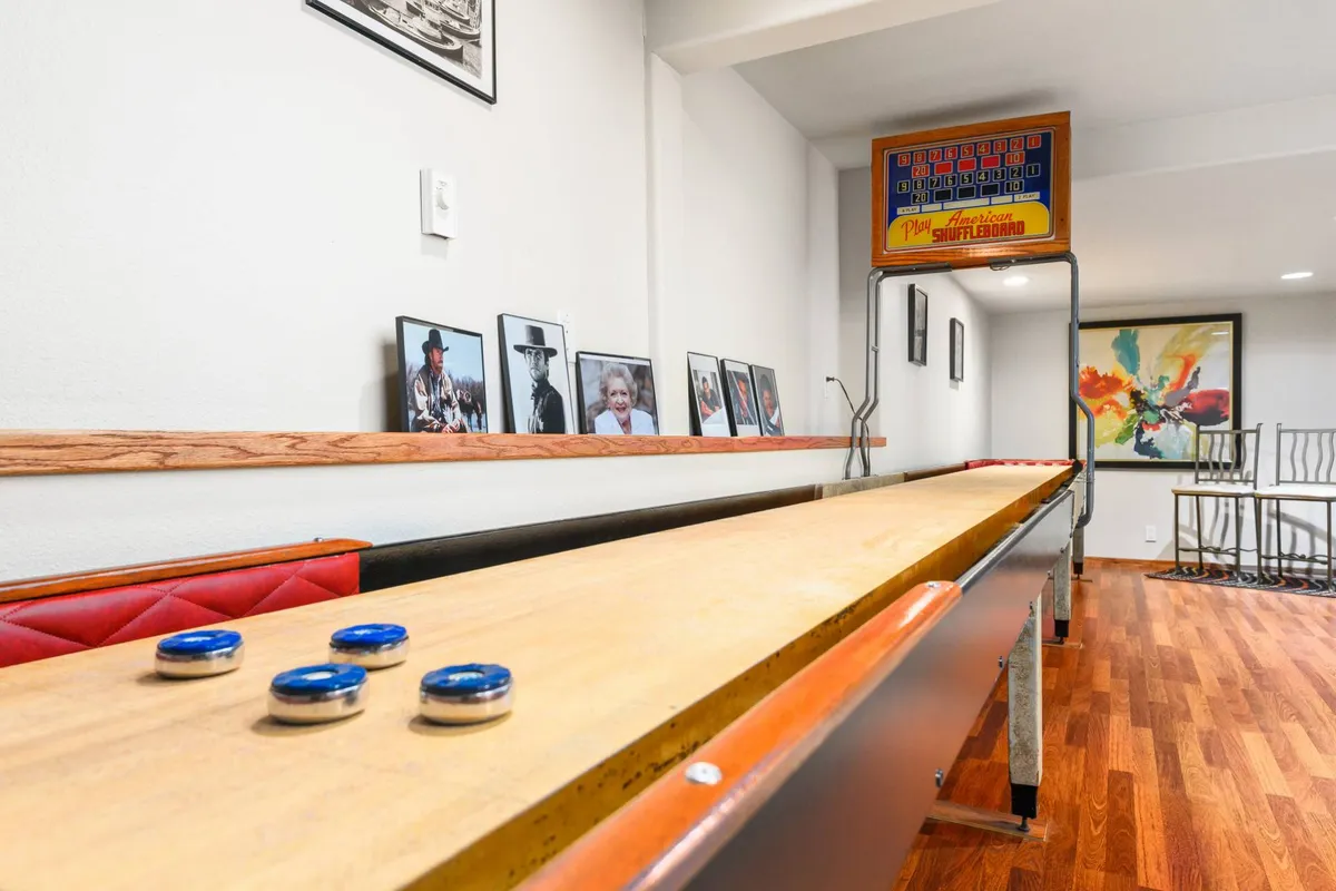 A vibrant game room featuring a shuffleboard table as the centerpiece, complemented by framed photographs and colorful artwork on the walls. Bar stools line the side, offering spectator seating and a social atmosphere ideal for friendly competitions and group gatherings.