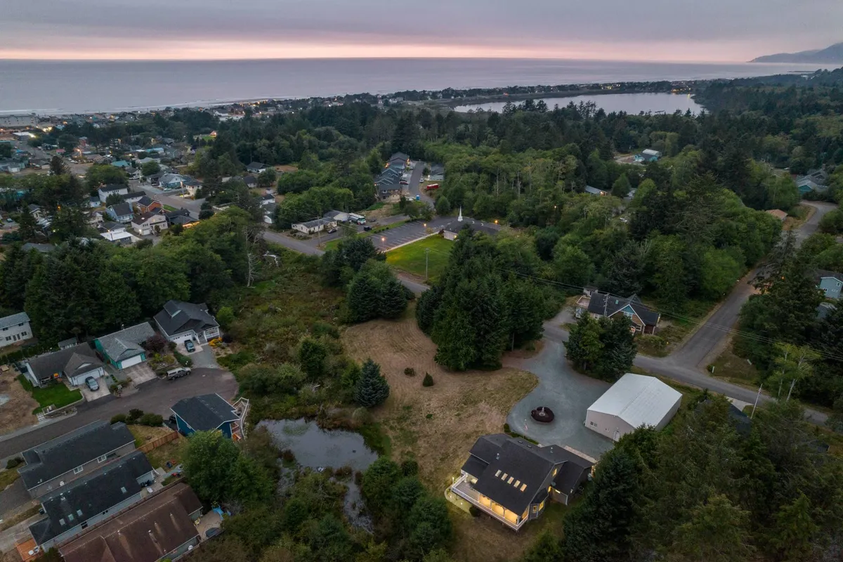 This image presents a serene ocean view framed by dense coastal greenery and hints of the nearby residential area. The expansive sky and shimmering water evoke the experience of being close to the beach and the natural beauty of the Oregon Coast.