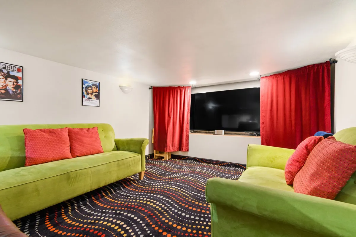 A plush, theater-style movie room with bright green sofas, red curtains, a large flat-screen TV, colorful carpet, and classic movie posters. The playful furnishings create a perfect atmosphere for movie marathons and group viewing.
