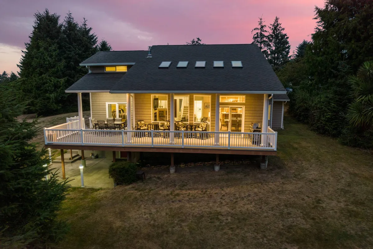 The image showcases the home's expansive deck nestled among lush trees, with large windows reflecting golden sunset light. Guests can be seen enjoying relaxed conversations, sipping coffee, and soaking in the tranquil wooded setting—all set against a peaceful backdrop that hints at both privacy and natural beauty.
