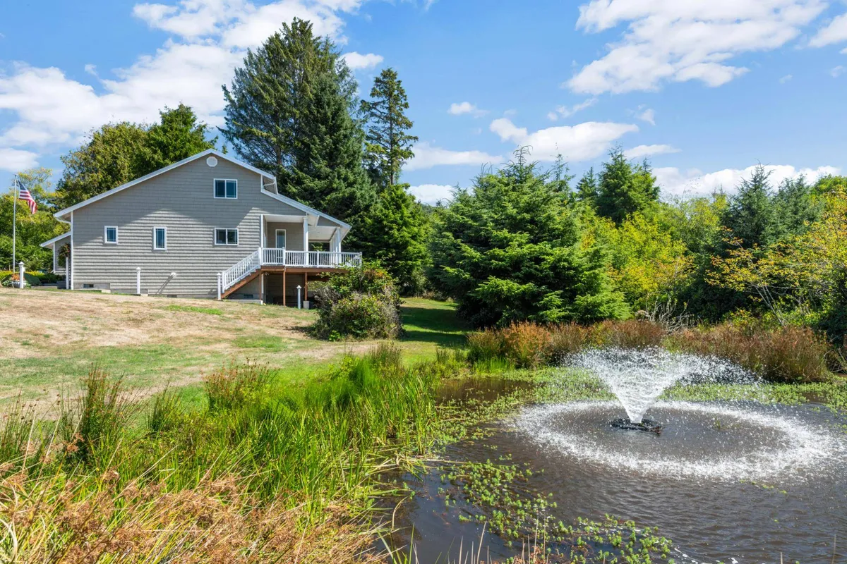 This exterior scene features the house set beside a serene pond with a fountain, surrounded by mature trees and lush greenery. The natural setting offers space for nature walks, outdoor relaxation, and team gatherings, with the porch visible for further outdoor enjoyment.