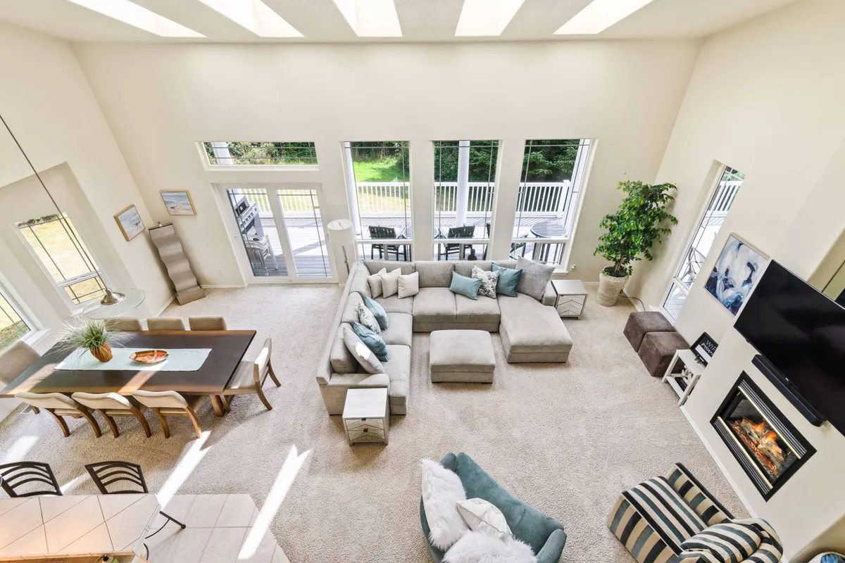 This image captures a large, open living space with a sectional sofa, a dining table, a fireplace, and expansive windows that let in abundant natural light. The arrangement of furniture encourages conversation and togetherness, and the clean, stylish décor makes the space both inviting and elegant.