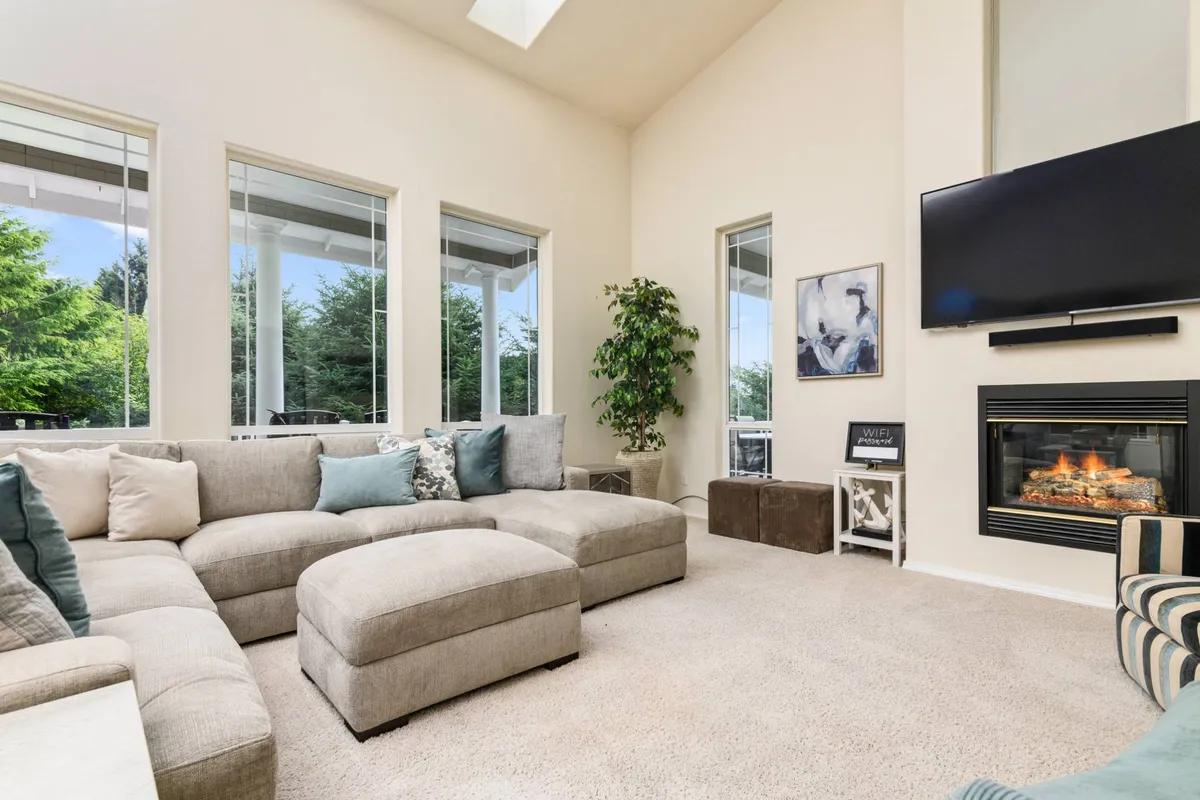 A spacious living room with a sectional sofa, fireplace, large windows, and indoor plants. Natural light floods the space, creating a warm and welcoming atmosphere ideal for relaxing after a day of adventures.