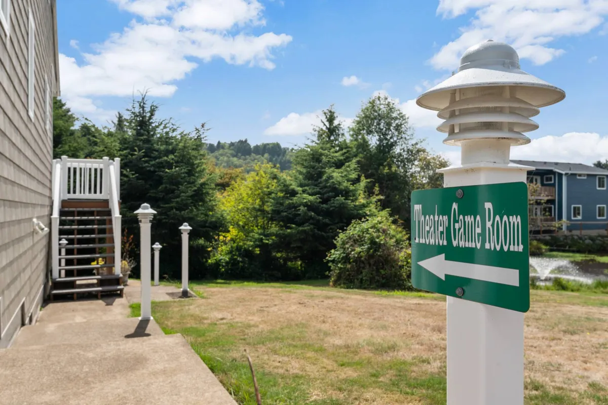 An exterior shot featuring the 'Theater Game Room' sign, stairs leading up to the entrance, and lush surrounding greenery. The scene hints at a dedicated entertainment outbuilding where guests gather for fun activities, all set against a playful and relaxed backdrop.