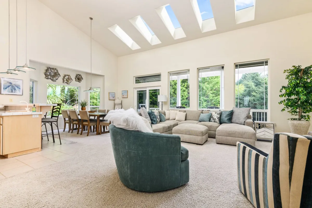 A wide-angle view of a comfortable interior featuring a large sectional sofa, a visible dining area, skylights flooding the room with natural light, and thriving indoor plants. The layout encourages interaction and adaptability for various family activities.